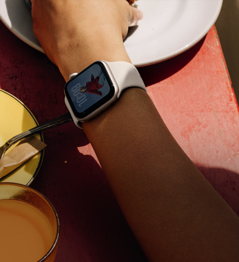 El brazo de una persona tomando comida de su desayuno, un Apple Watch SE 3 en la muñeca, caja de aluminio blanco estelar, pantalla siempre activa, carátula con el retrato de una persona, correa deportiva blanco estelar.