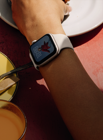 El brazo de una persona tomando comida de su desayuno, un Apple Watch SE 3 en la muñeca, caja de aluminio blanco estelar, pantalla siempre activa, carátula con el retrato de una persona, correa deportiva blanco estelar.