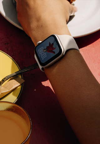 El brazo de una persona tomando comida de su desayuno, un Apple Watch SE 3 en la muñeca, caja de aluminio blanco estelar, pantalla siempre activa, carátula con el retrato de una persona, correa deportiva blanco estelar.